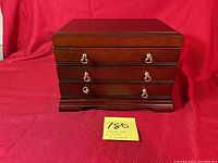 Closed dark polished wood jewelry box with three drawers and gold-tone handles on a red background.