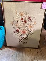 Full view of the large framed embroidery with floral bouquet in basket on tan background.