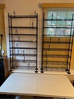 Two black metal wire CD racks side by side on a table, under a window, showing full height and shelf details
