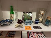 Shelf display of all miscellaneous knickknacks including Johnson Space Center dish, dragon turtle figurine, colonial map paperweight, glass bottles and jars with marbles and seashells, small ceramic dishes and decorative items.