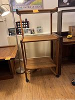 Front view of solid oak multi-purpose cart with three shelves and caster wheels, showing the top shelf with raised edges and the overall design and wood finish.