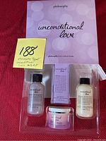 Top-view photo of the Philosophy Unconditional Love small body care kit with shower gel, body cream, spray fragrance, and brand card on a red cloth background.