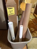 Several rolled posters in brown and white tubes standing inside a beige plastic container. The container has a sign indicating '167 Posters'.