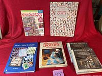 Five quilting and crochet books laid out on red fabric backdrop showing front covers including World War II Quilts, Carry-Along Crochet, Gathering by Artists American Quilts, Sew Scrappy, and Hobo Quilts duplicate.