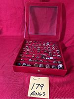 Open red faux leather case displaying multiple rows of assorted women's rings on red velvet lining, note reading '179 Rings' visible