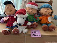 Four Peanuts Gang soft toys including Charlie Brown, Snoopy, Linus and a fourth character, all about 10 inches tall, seated on a wooden surface near a curtain.