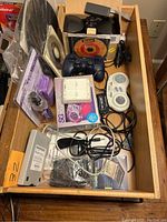 Top-down view of drawer containing various vintage gaming and computer accessories including game controllers, DS cartridge case, speakers, mouse, microphone, cleaning kits, and other gadgets.