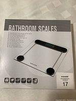 Photo of front of the box showing the image of the transparent electronic bathroom scale with black circular sensors and digital display.