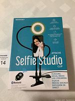 Front view of the packaging showing the selfie studio clip with ring light around phone holder and clamp base. Bluetooth logo visible.
