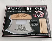 Front view of the new Alaska Ulu Knife in original packaging showing wood handle, stainless steel curved blade etched with bear design, and stand.