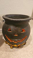 Front view of the cauldron pumpkin showing carved triangular eyes, nose and a jagged smile with orange and yellow paint inside carved areas.