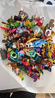Top-down image showing a pile of assorted vintage character toys including animals, cartoon characters, and accessories. Various colorful plastic figures packed closely together on a white backdrop.