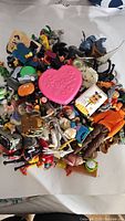 Pile of assorted vintage toy figures and small toys including pink Barbie heart container, various character figurines and small vehicles.