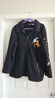 Full front photo of black fleece zip-up jacket hanging on a hanger showing embroidered Pooh and Eeyore on chest and some leaves embroidery on sleeve.