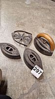 Four antique sad irons arranged on a concrete floor, one with wooden handle and three flat irons.