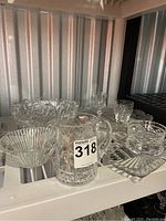 Wide angle photo showing assorted crystal including large pitcher, bowls, plates, and napkin rings on white shelf under storage lighting.