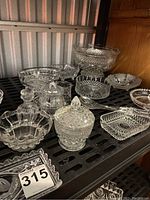Multiple assorted clear crystal glassware pieces including bowls, jam pot, and small dishes arranged on a black shelf