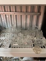 Full view of assorted crystal and vintage glassware on a metal shelf including small glasses, jam pots, salt and pepper shakers and a McKee glass salad bowl
