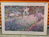 Full frontal image of the framed Claude Monet print showing the garden scene with purple flowers, the gold-colored frame around it, and the small crack detail on the glass at the lower right.