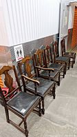 Six vintage wooden dining chairs and one captain's chair arranged in a row against a wall. Chairs have carved wooden backrests and black cushioned seats.