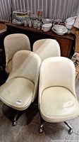 Set of four cream-colored mid-century vinyl dining chairs with castors, arranged in a group showing seats and backs.