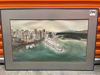 Framed watercolor painting showing a cityscape of buildings along a waterfront with hills in the background. The scene is rendered in soft colors and visible through a glass frame.