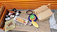 Overview of wicker picnic basket, coffee cups, rolling pins, glass baking dish, small plates and collectors tins on wooden bench
