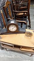 Photo of two time pieces placed on a wooden table with wooden chairs in background. Larger wooden mantle clock with arched shape and smaller round Westclox alarm clock are visible.