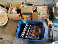 Blue plastic tote filled with upright vinyl records, and surrounding cardboard boxes