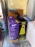 Photo showing unopened boxed Bushnell and Optex binoculars stacked side by side.