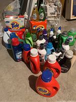 Photo showing a wide view of all cleaning supplies arranged on the floor including multiple Tide orange bottles, Downy, Clorox, Palmolive, Greenworks, Jergens, and Listerine products.
