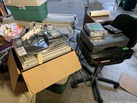 Various electronic components including turntable in cardboard box, audio speakers, and stacked video VCRs on a chair.