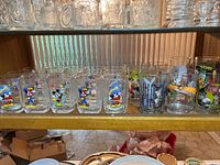 Shelf holding 13 various collectible glasses featuring Disney characters and movie designs.