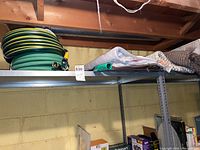 Shelf holding two Yardworks garden hoses with tags intact, along with a green watering wand and several rolled mats of different textures and colors.