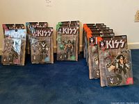 Nine unopened Kiss action figures in original packaging displayed on a blue carpet, showing Paul Stanley, Ace Frehley, Peter Criss, and Gene Simmons figures.