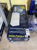 Two unopened boxes of Stack-A-Shelf 3 Drawer Unit organizers stacked on concrete floor in a basement area near other items.