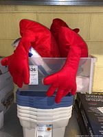 Two red heart-shaped pillows with extended arms placed inside a clear plastic bin on top of stacked Rubbermaid Roughneck bins with blue and white lids.