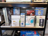 Shelf with multiple boxed kitchen and personal care appliances including Silk'n Glide Hair Removal device, Salton coffee maker, springform pans, ceramic cannister, and others.
