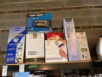 Photo of boxed items including Dr. Scholl's Comfort Plus Foot Bath, Black & Decker toaster oven, Melitta coffee maker, and other boxed personal care and small appliance products on shelving against a brick wall.