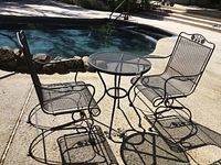 Outdoor photo showing black metal bistro set with two rocking chairs and a round mesh table beside a pool.