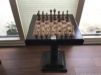 Top front view of wooden chess board with chess pieces set up, mounted on pedestal base.