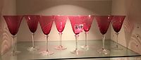 Seven cranberry-colored glass goblets arranged in a row, showing their conical shape and translucent pinkish-red tint with clear stems.