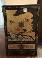 Front view of cabinet showing black lacquer finish with gilt crane, red sun and pine tree motif, brass latch
