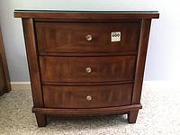 Front view of a Somerton nightstand showing three curved front drawers with metal round knobs and a glass top surface.