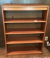Front view of the teak bookcase showing four shelves with visible wear and wood grain.