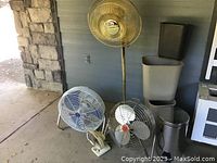 Overview showing four indoor fans of different sizes and styles, with four plastic trash containers and one metal trash can stacked to the side.