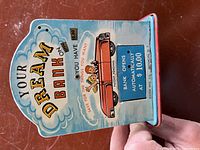 Front side of the bank showing 'YOUR DREAM BANK' text, red convertible car illustration, and instruction text 'SAVE FOR THE THINGS YOU WANT, BANK OPENS AUTOMATICALLY AT $10.00'