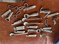 Lot of assorted vintage metal bottle openers laid out on a surface, showing different types including openers with knives, corkscrews, and spoons.