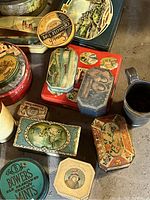 Photo showing an assortment of vintage tins including green peppermint tin, floral octagonal tin, scenic and portrait tins in various shapes and colors.