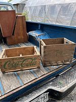 Two wooden crates in the bed of a blue truck, one Coca-Cola crate and one Pepsi crate, both in solid condition.
